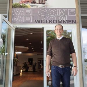 Lars Busse Geschäftsführer von Fachwerk Küchen steht vor dem Eingang seines Küchenstudios. Über ihn weißt ein Schild Willkommen auf dein Eingang hin.